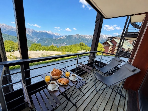 Ein Balkon mit Frühstückstisch, ausgestattet mit Croissants, Saft und Kaffee, mit Blick auf eine Berglandschaft.