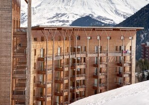 Ein mehrgeschossiges Gebäude mit Balkonen, einer Holzfassade und einem Bergpanorama.