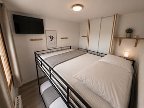 Un lit simple avec une tête de lit, une table de chevet avec une lampe, un téléviseur fixé au mur, un tableau encadré et une petite étagère avec une plante.
