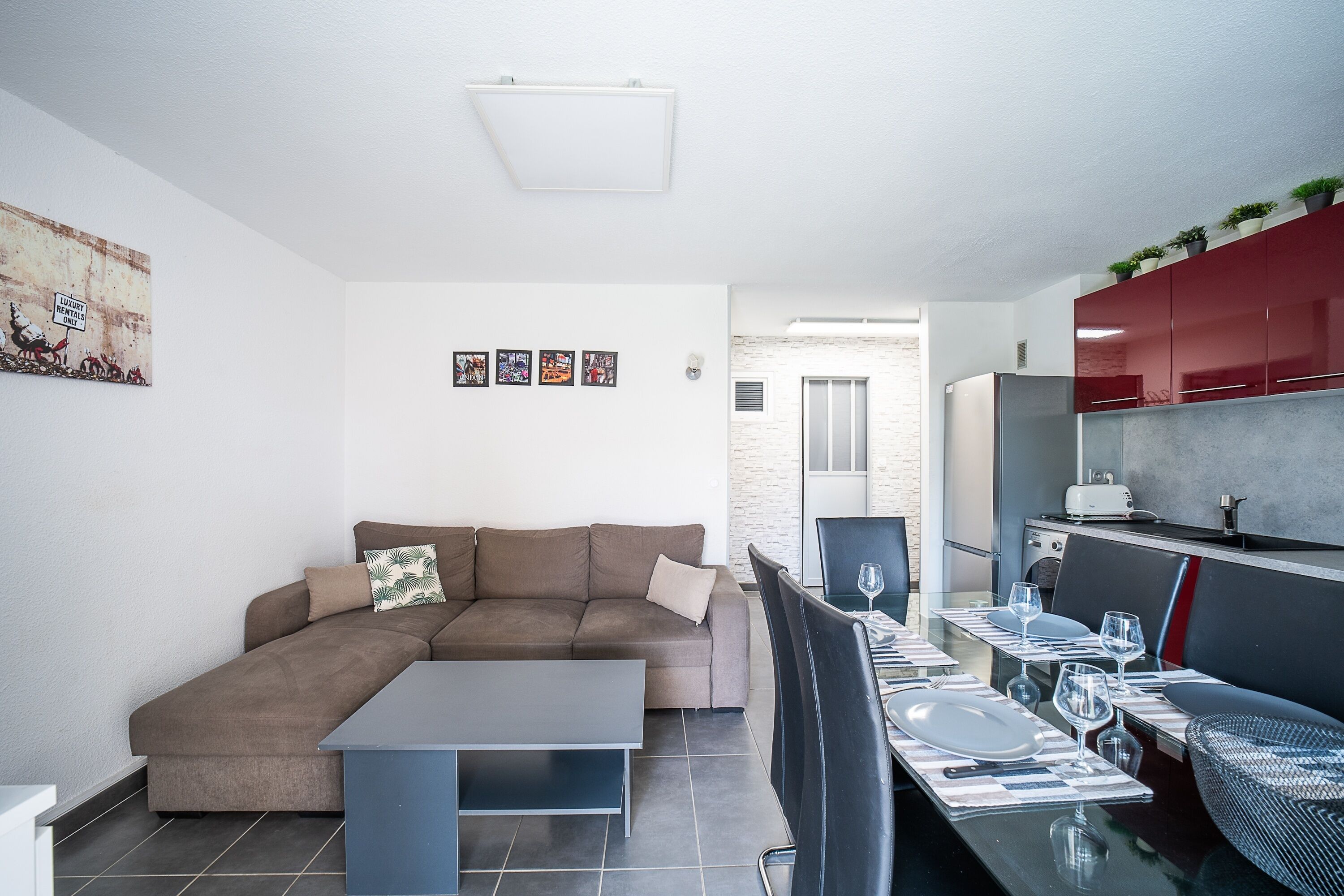 A modern living room with a sectional sofa, a dining table set for four, and a kitchen area with red cabinets and a built-in oven.