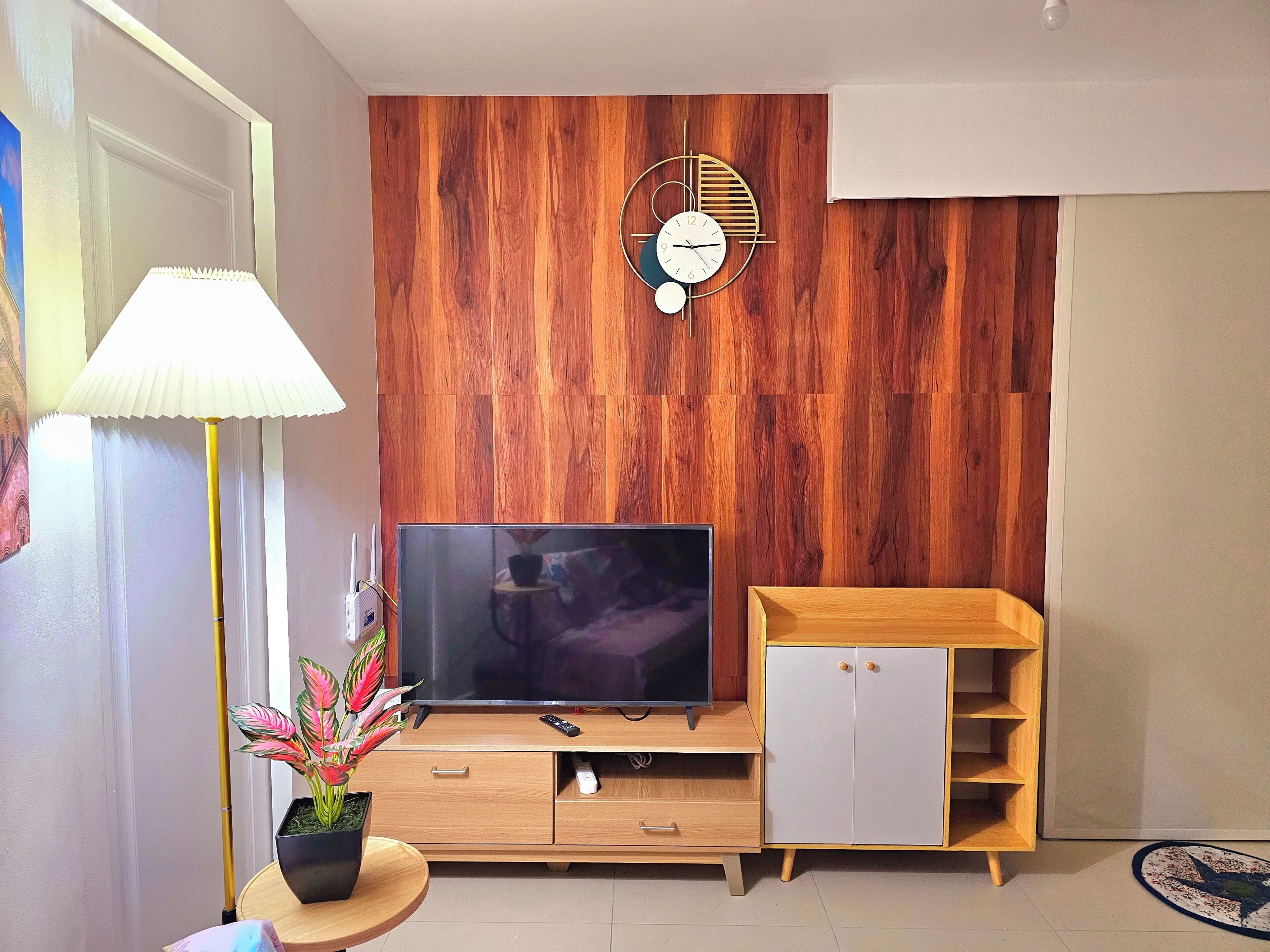 A modern living room with a wooden wall panel, a flat-screen TV, a wooden cabinet, a lamp, and a clock.