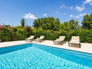 Une piscine entourée de quatre chaises longues, le tout baigné d’une végétation luxuriante et surplombé d’un ciel bleu azur.