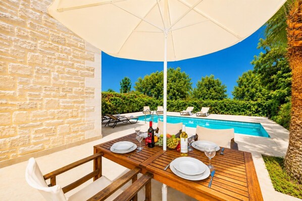 Une table en bois, avec de l’assiette blanche et des verres à vin, un parasol blanc, et une piscine avec des chaises longues en arrière-plan.