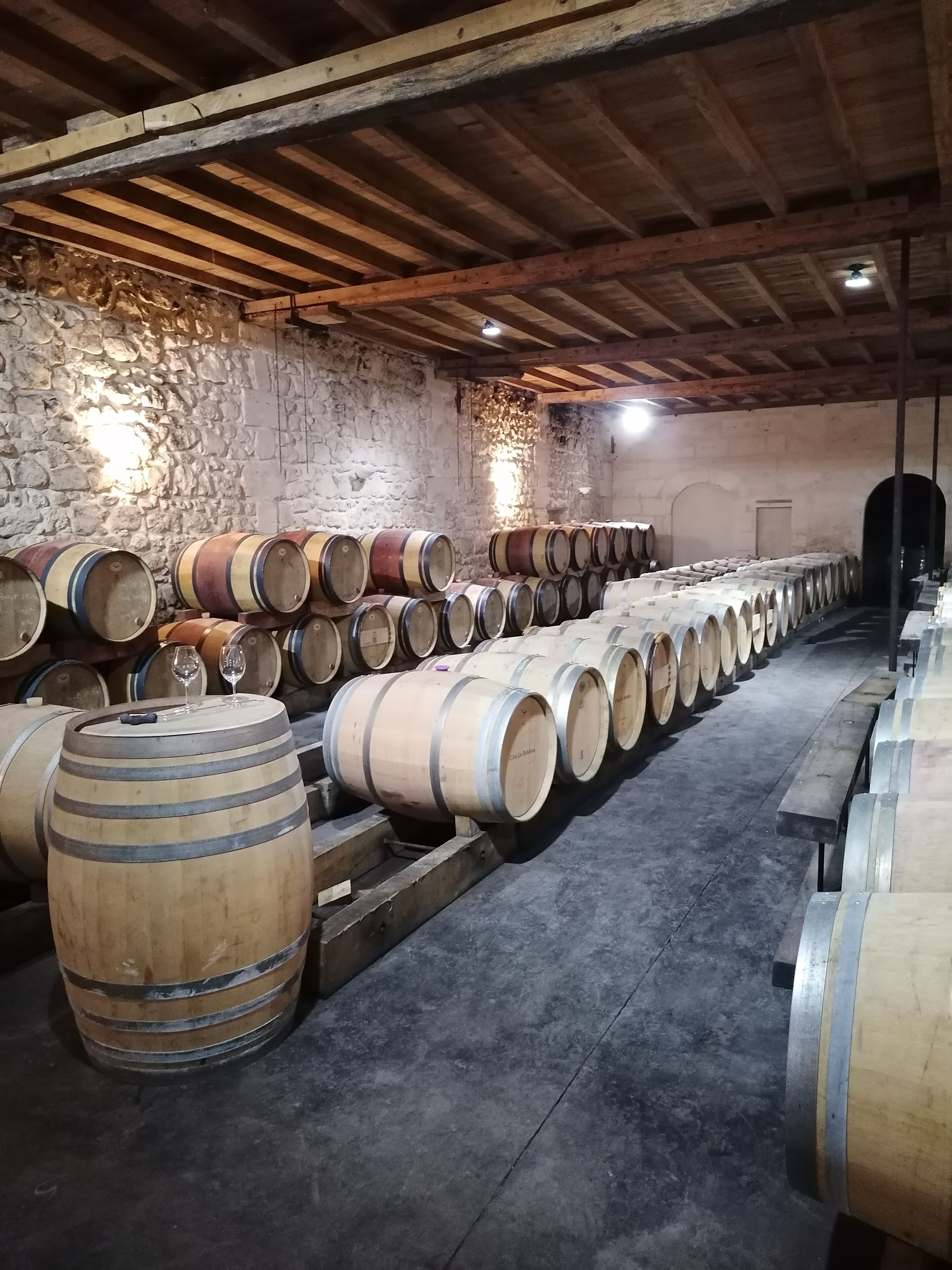 Une cave avec des barriques en bois disposées en rangées, un mur en pierre et un plafond en bois.