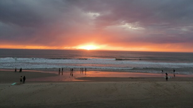 Una playa con gente caminando y practicando surf, y el océano al fondo.
