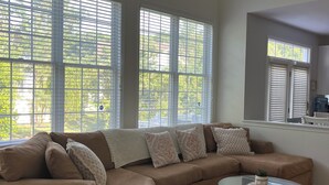 A living room with a beige sectional sofa, a glass coffee table, and large windows with blinds.