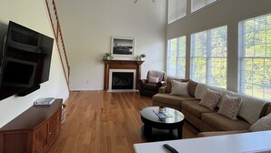 A spacious living room with hardwood floors, a fireplace, a ceiling fan, and large windows.
