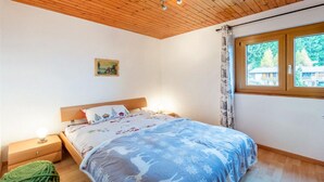A bedroom with a wooden ceiling, a bed with a blue and white patterned bedspread, a nightstand with a lamp, and a window with a view of a house. - Chalet in Les Collons by Ski Slope (Vex)