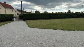 Un camino de grava conduce a una casa con tejado rojo, rodeada de un seto cuidado y un césped.