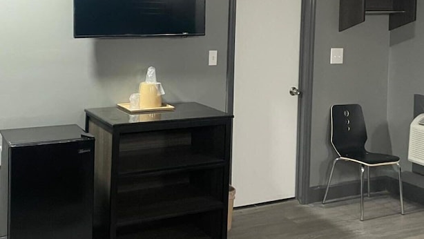 A room with a flat-screen TV mounted on the wall, a small black cabinet, a door, a chair, and a patterned rug on the floor. - Travelers Rest Motel (Birmingham)