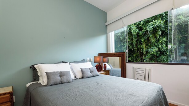 A bedroom with a bed, a nightstand, a mirror, and a window with a view of greenery. - BR-96 (Rio de Janeiro)