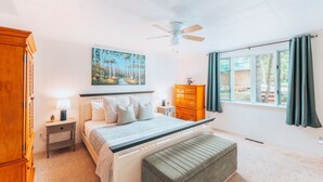 Une chambre à coucher comprenant un lit, une table de chevet, une commode, une fenêtre avec des rideaux, un ventilateur de plafond et un tableau accroché au mur.
