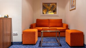 Um quarto pequeno e acolhedor com um sofá laranja, dois pufes iguais e uma mesa de centro transparente. Uma janela com vista para a paisagem urbana está acima do sofá.