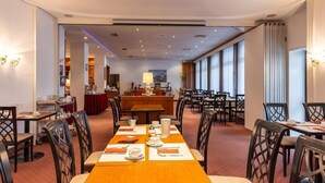 Uma sala de jantar de hotel com uma mesa comprida posta para refeição, rodeada por cadeiras, e uma área de bar ao fundo.