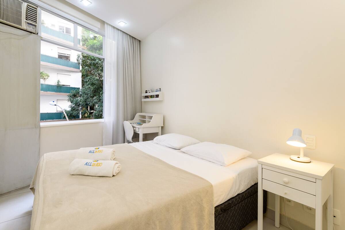 A hotel room with a bed, a desk, a chair, a nightstand, and a window with curtains.