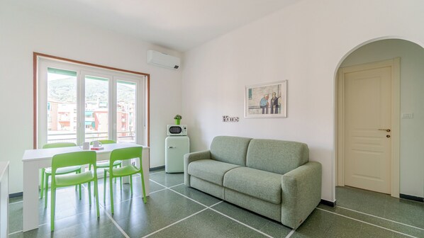 Ein Zimmer mit einem grünen Sofa, einem Esstisch mit Stühlen, einer kleinen weißen Kommode und einem Bild an der Wand.