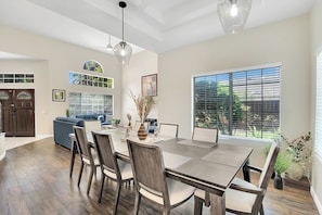 A dining area with a large table, chairs, and pendant lights. There is a kitchen area with a double door and a window with blinds.