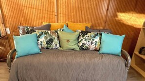 Ein Zimmer mit Holzwänden, einem grauen Schlafsofa, mehreren Kissen und einem kleinen Holzregal.