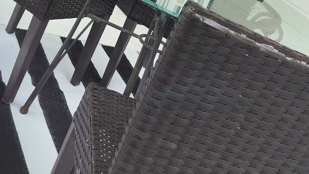 Een overdekt terras met rieten stoelen, een glazen tafel, een witte lantaarn en een zwart gekaderd raam.
