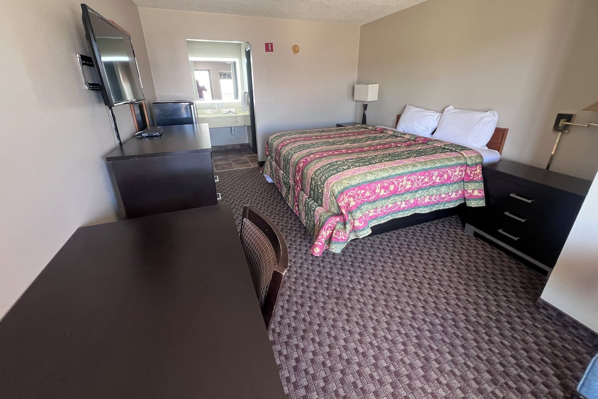 A hotel room with a bed, a desk with a chair, a TV, and a bathroom visible through an open door.