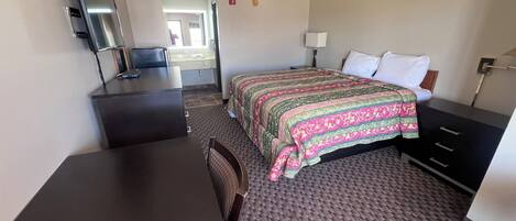 A hotel room with a bed, a desk with a chair, a TV, and a bathroom visible through an open door.