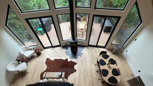 Un intérieur moderne avec un plancher en bois, un tapis central, un canapé noir, une table ronde avec des chaises et de grandes fenêtres donnant sur une terrasse extérieure.