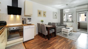 Een moderne keuken met een ingebouwde oven, vaatwasser en koffieapparaat. Er is een eethoek met een tafel en stoelen, en een zithoek met een bank en een klein witte bijzettafeltje.