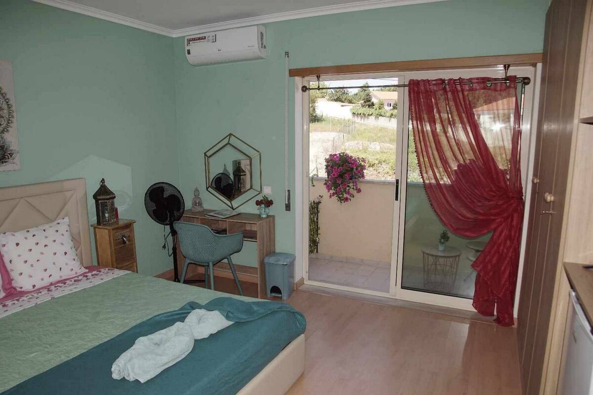 A bedroom with a bed, a desk, a chair, a small table, a heater, and a sliding glass door to a balcony.