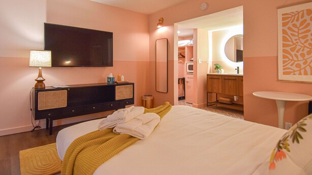 Une chambre d’hôtel comprenant un lit, une commode, un téléviseur, un miroir, un petit miroir rond, une petite table et une salle de bain visible à travers une arche.