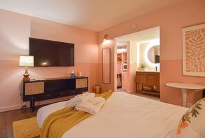 Une chambre d’hôtel comprenant un lit, une commode, un téléviseur, un miroir, un petit miroir rond, une petite table et une salle de bain visible à travers une arche.