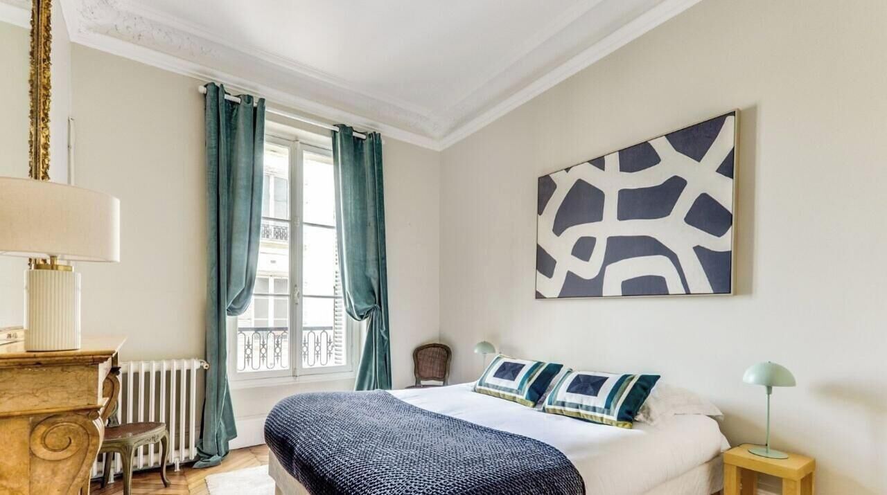 Une chambre à coucher comprenant un lit, une table de chevet, une commode, une fenêtre avec des rideaux et une œuvre d’art accrochée au mur.