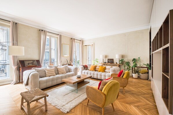A spacious living room with a sofa set, a coffee table, and a wicker chair. There are large windows with curtains, a piano, and a bookshelf.