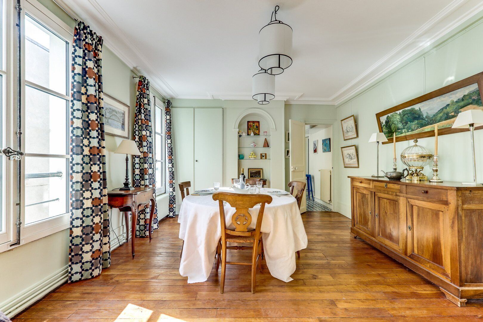 A dining area with a wooden table set for four, a wooden cabinet with decorative items, and a large window with patterned curtains.