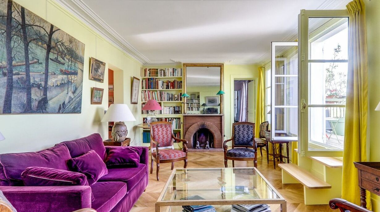 A living room with a purple sofa, a fireplace, bookshelves, and a large window with curtains.