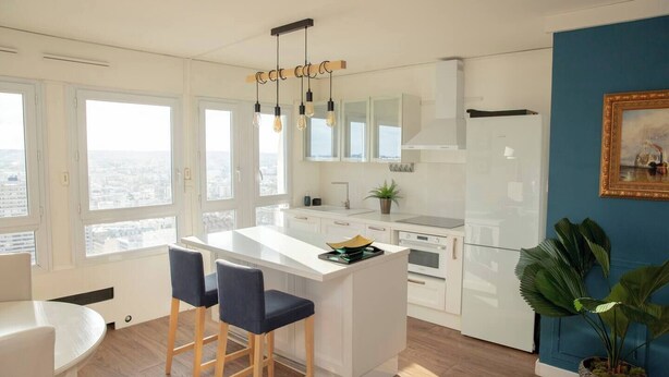 Eine moderne Küche mit weißen Schränken, einer zentralen Kochinsel mit Barhocker und Blick auf die Stadt durch große Fenster.
