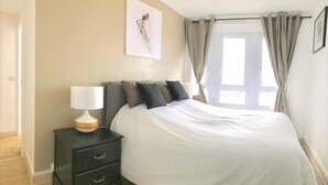 Ein Schlafzimmer mit Bett, Nachttisch, Lampe und einem Fenster mit Vorhängen.
