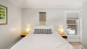 Ein ordentlich eingerichtetes Schlafzimmer mit einem großen Bett, zwei Nachttischen mit Lampen, einer Wanddeko aus Makramee und einem Fenster mit Blick auf eine verschneite Landschaft.