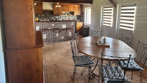 A dining area with a round table and chairs, a kitchen in the background, and a window with blinds.