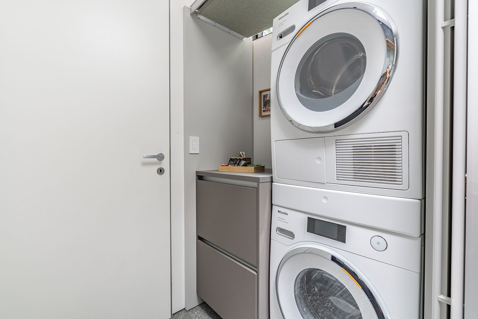Una sala de bugaderia amb piles de rentadores i assecadores, una taula petita amb objectes i una porta.