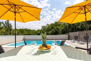 A poolside table with yellow umbrellas, a pineapple, bananas, an orange, and wine glasses. - Charming holiday house Aurora with private pool (Gabonjin (Krk))