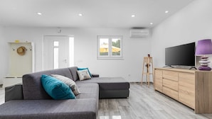 Un salon moderne avec un canapé d’angle gris, un meuble en bois sur lequel est posée une télévision à écran plat, et une fenêtre donnant sur un parapluie jaune.