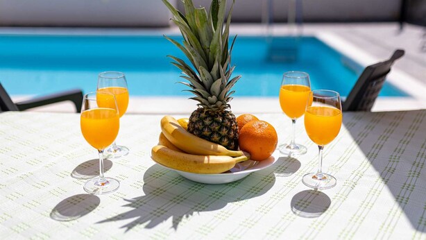 Une table avec une assiette de fruits et des verres de jus d’orange, près d’une piscine.