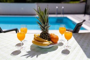 A table with a plate of fruits and glasses of orange juice by a pool.