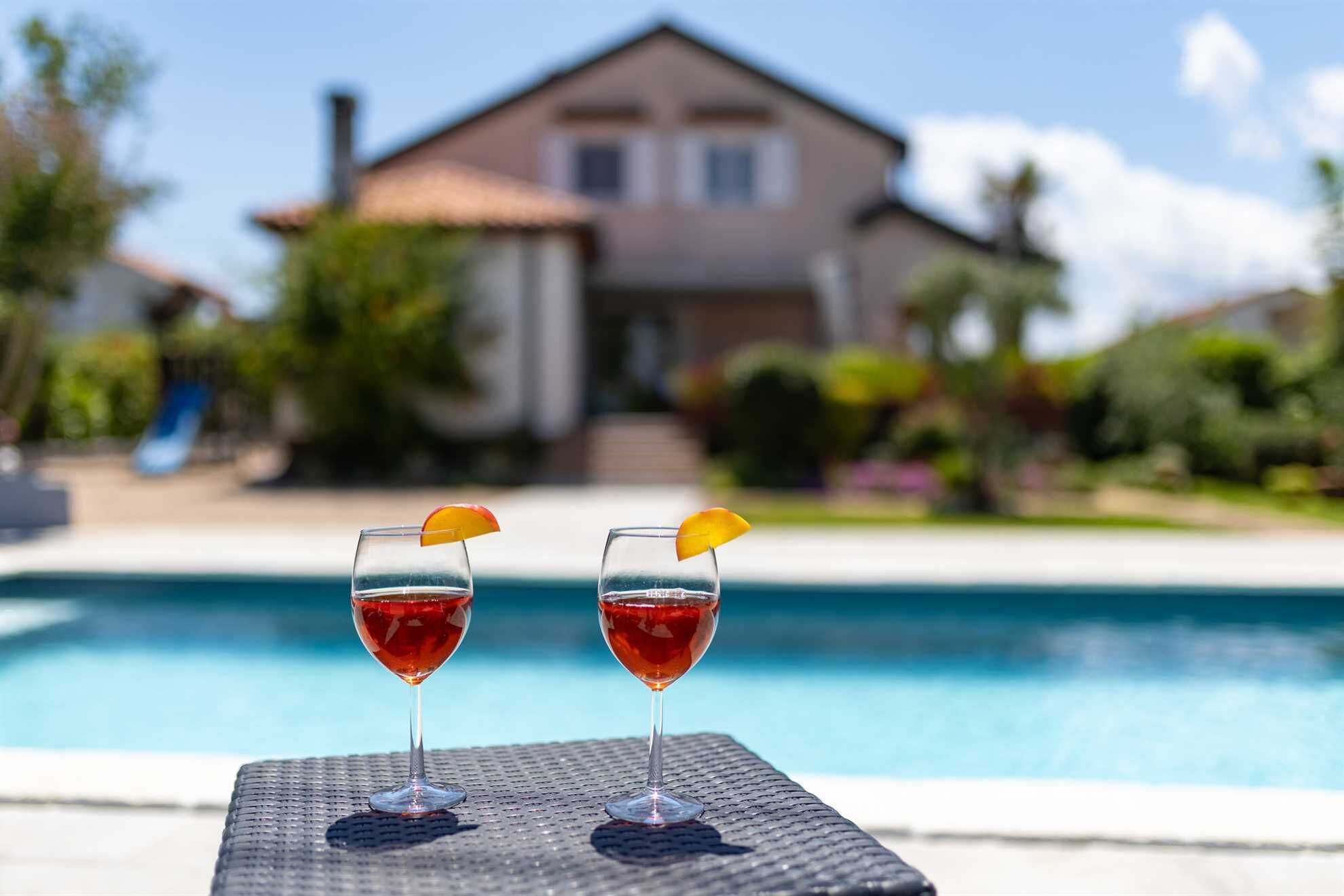 Two glasses of red beverage with orange slices on a poolside table with a house in the background.