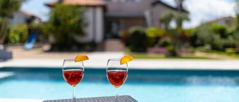 Two glasses of red beverage with orange slices on a poolside table with a house in the background.