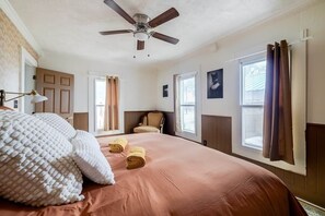 Un dormitorio con una cama grande, ventilador de techo, ventana con cortinas y una silla.
