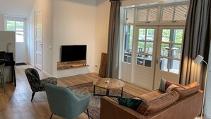 A living room with a flat-screen TV mounted on a wooden shelf, a brown sofa, a blue armchair, and a small round table. - Apartment in Wageningen Near Rhine River (Elst)