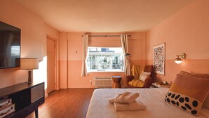A hotel room with a bed, a TV, a window with curtains, a chair, and a small table with a lamp. - Mid-Century Themed Hotel Room in Fayetteville, AR - The Clementine on College #5 (Fayetteville)
