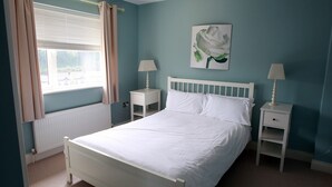 A bedroom with a bed, two bedside tables, a window with curtains, a radiator, and a wall-mounted painting. - Glen Ard Holiday Home no. 1 (Dunmore East)