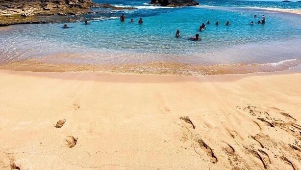 Ein Strand mit klarem, blauem Wasser, Menschen beim Schwimmen und Fußabdrücken im Sand.
